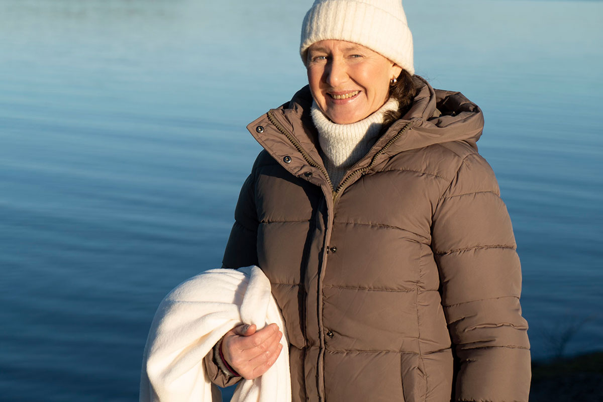 Vibeke står ved vannet en kald vinterdag i boblejakke og med håndkle, klar for å isbade